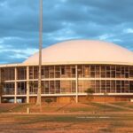 Prédio da CLDF em Brasília com bandeira LGBTQIAPN+ ao fundo, sob céu nublado, destacando programa Reconhecer e críticas por atraso.