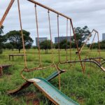 Parquinho público negligenciado em Brasília com equipamentos quebrados e mato alto, representando falha de manutenção do GDF.