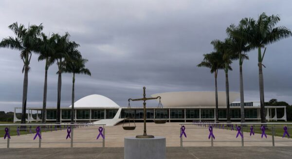 Edifício da CLDF em Brasília com símbolos de impunidade na violência de gênero, como fitas roxas e balança desequilibrada.