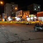 Cena de crime em rua de Taguatinga com fita policial, moto caída e viaturas, representando homicídio por atirador em moto.